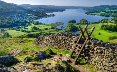 A morning shot of Lake Windermere and surrounding hills with a stone wall and stile in the foreground.