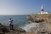 Portland Bill lighthouse is a historic working lighthouse on the Isle of Portland's most southerly headland. It is painted red and white. Two people, a couple standing on the rocks on the shoreline, with the lighthouse in the background.