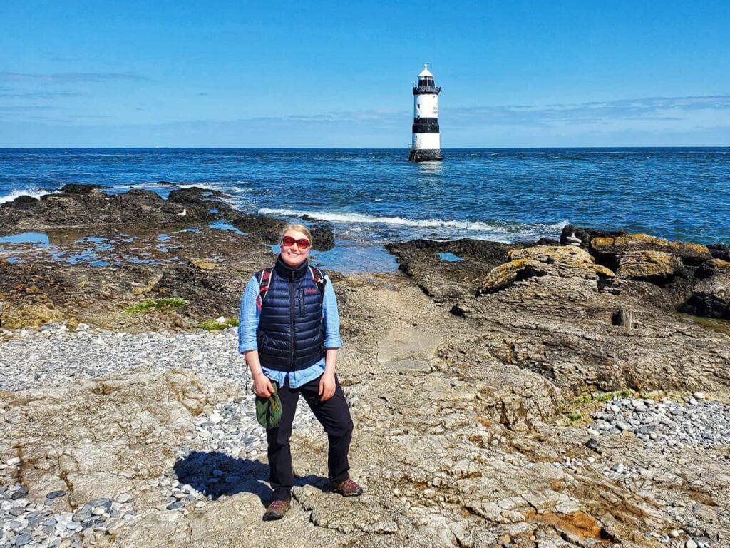 Sine standing by Trwyn Du Lighthouse on the Anglesey Coastal Path, with waves crashing on the rocky shore