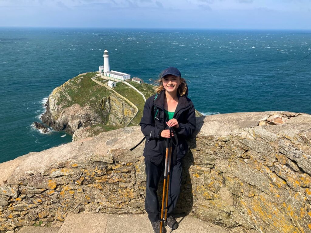 Walker standing near the cliff edge with South Stack Lighthouse and the Irish Sea in the background.