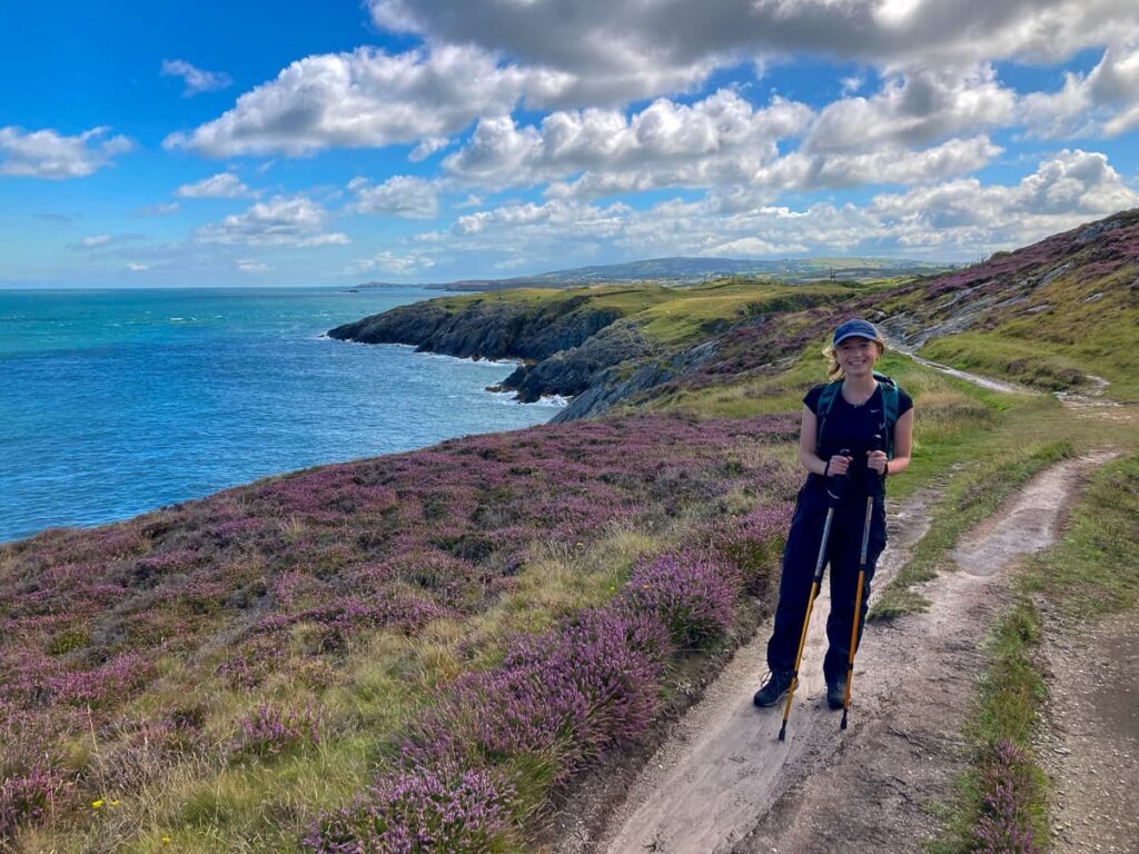Walker enjoying purple heather-covered paths near Trwyn Myn with coastal cliffs stretching ahead.