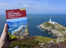 Holding a Wales Coast Path guidebook with South Stack Lighthouse and turquoise sea in the distance.