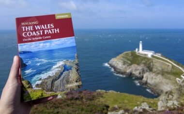Holding a Wales Coast Path guidebook with South Stack Lighthouse and turquoise sea in the distance.