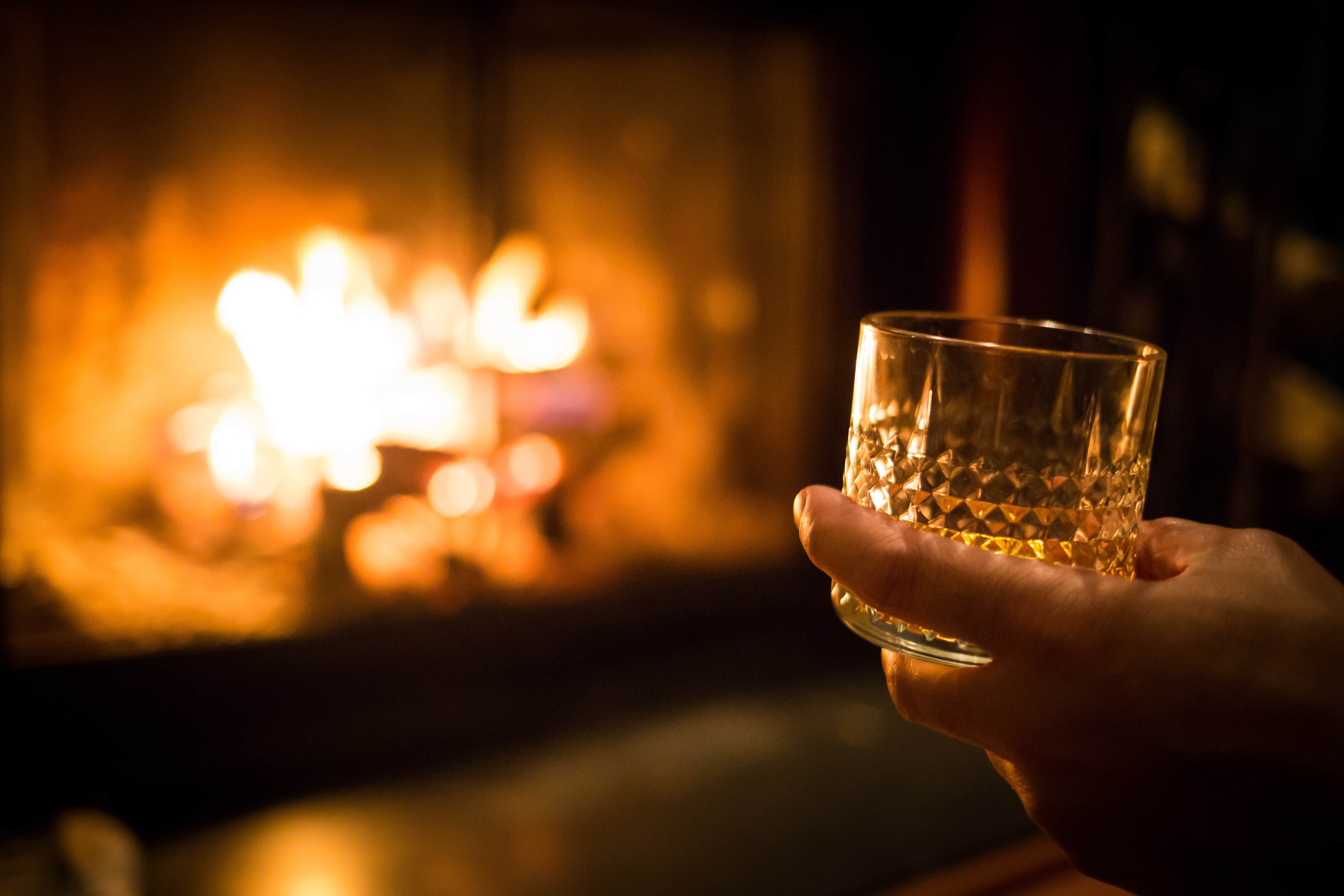Hand holding a whisky glass by a fireplace, symbolising cosy winter travel in Scotland